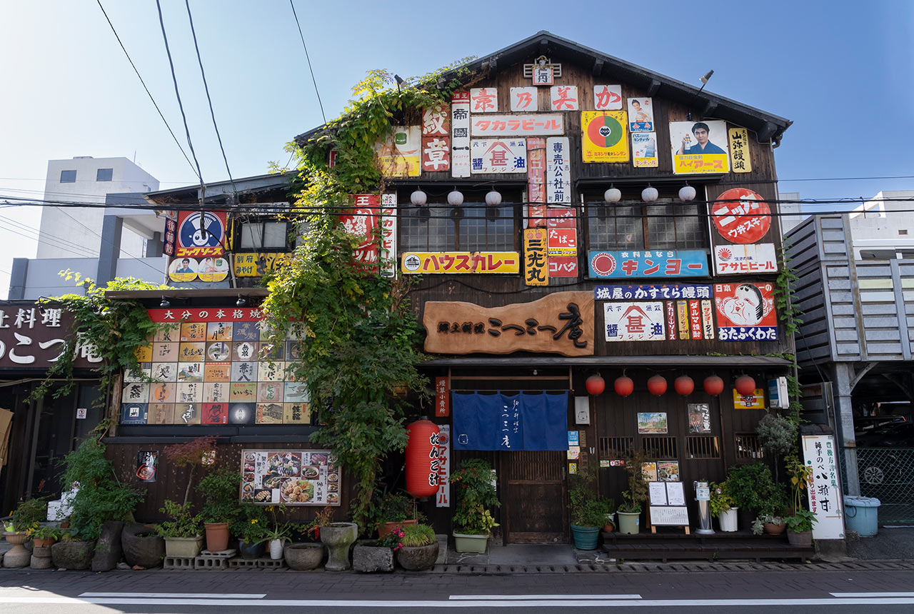 〈郷土料理 こつこつ庵〉の外観。壁一面に看板や焼酎ラベルが飾られている